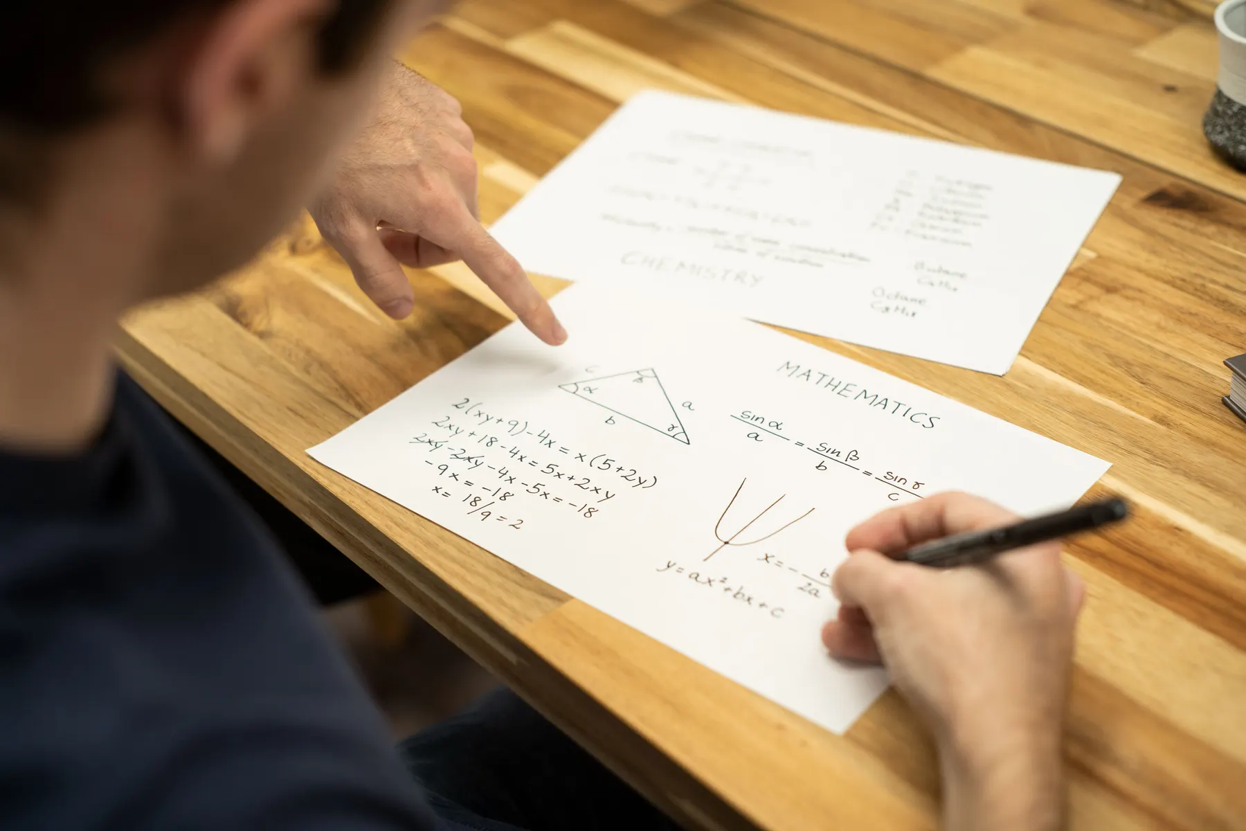 A tutor pointing at a worked algebra problem on a sheet of paper beside a student